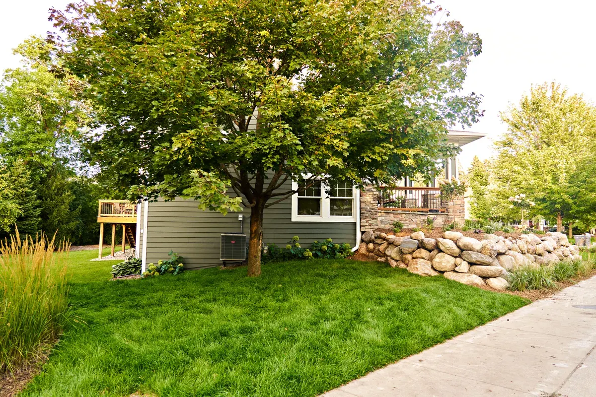 Boulder retaining wall with ornamental grasses along a sidewalk beside a gray-sided home