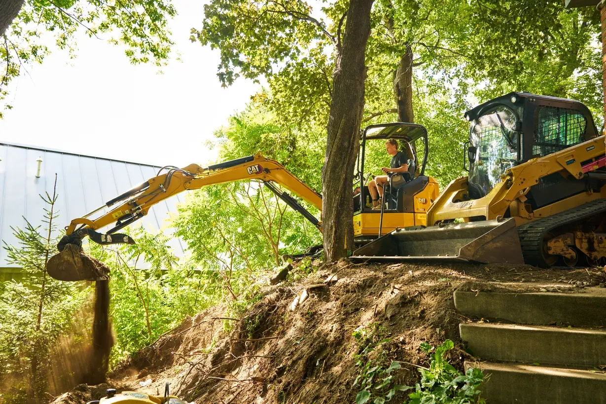 Excavator digging into a wooded hillside beside a track loader