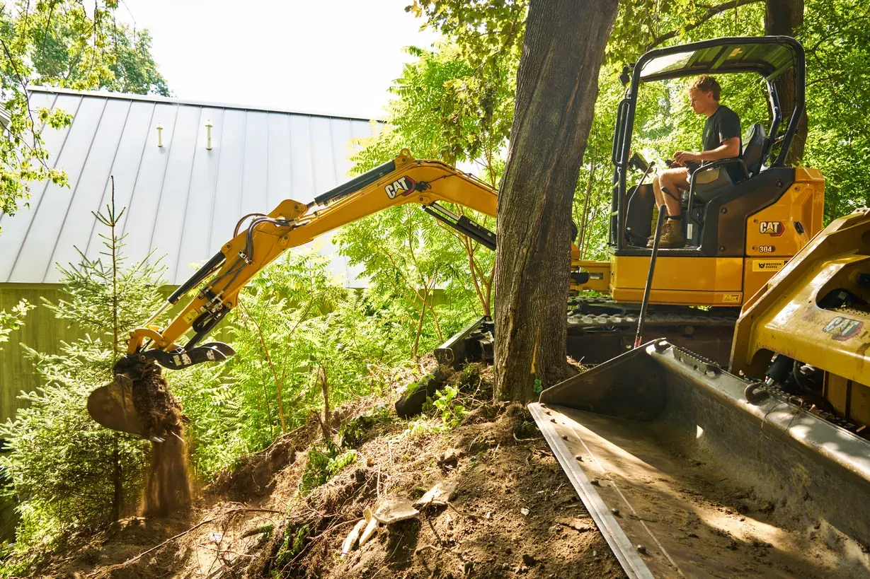 Excavator digging into a wooded hillside beside a track loader